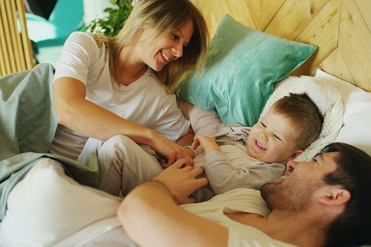 Parents et enfant sourient dans le lit, moment paisible et complice de vacances.