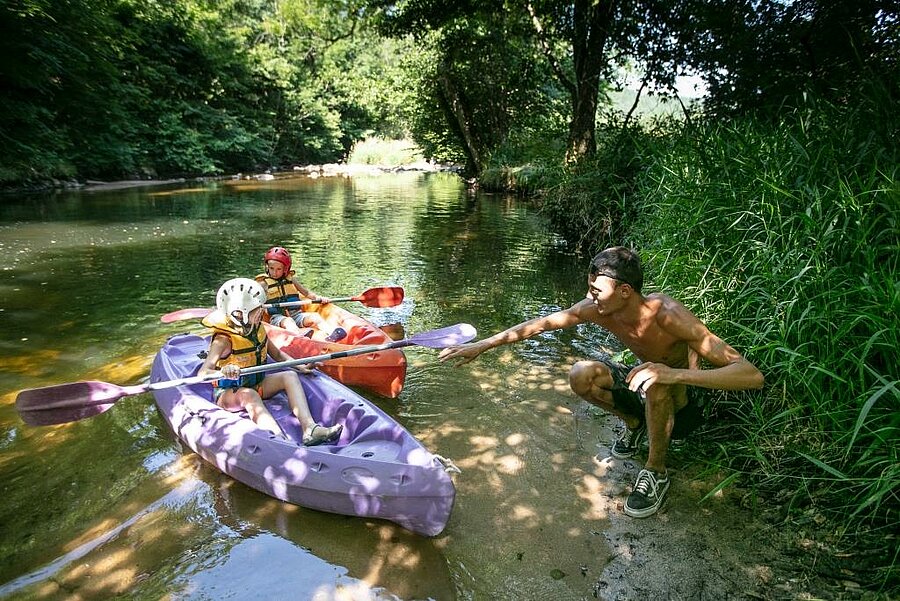 Enfants en kayak sur une rivière près d’un camping bord de lac, moment convivial entre nature, baignade et loisirs nautiques.