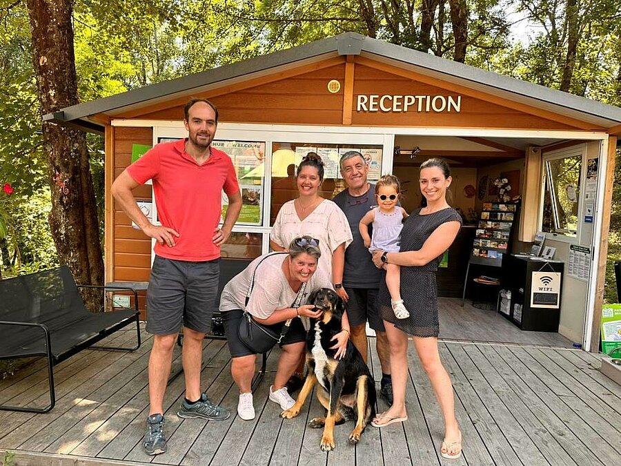 Les propriétaires d'un campings posent souriants devant la réception, accompagnés de vacanciers et de leur chien, dans un cadre naturel et convivial.