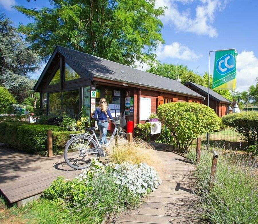 Entrée accueillante et fleurie d’un camping dans le Loiret, avec une cycliste près de l’accueil, illustrant l’atmosphère chaleureuse et la proximité avec les activités de plein air comme le vélo.