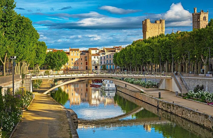 Vue sur le canal de la Robine à Narbonne, bordé d’arbres et dominé par la tour médiévale, près d’un camping nature.