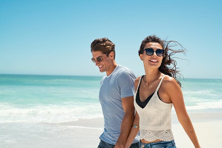 Couple main dans la main sur une plage en été, ciel bleu et océan, idéal pour un séjour à deux au grand air.