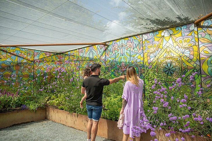 La galerie des papillons © Terra Botanica Des visiteurs admirent, à l'abri de la pluie, des fleurs violettes devant des murs aux motifs végétaux colorés.
