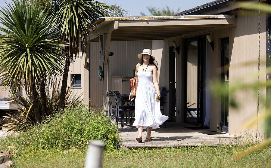 Femme en robe blanche sortant d’un chalet moderne avec terrasse ombragée, dans un camping près de Toulouse entouré de verdure.
