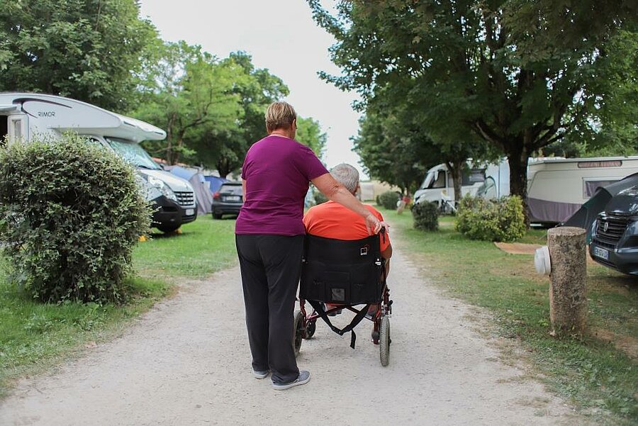 Femme accompagnant un homme en fauteuil roulant sur une allée de camping arborée, proche des emplacements PMR.