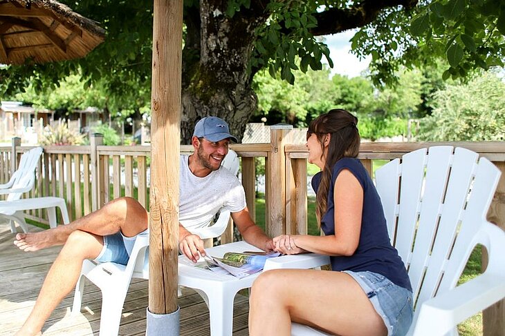 Moment détente en terrasse pour un early booking couple Couple souriant assis sur une terrasse ombragée en camping, scène de détente illustrant l’early booking couple.