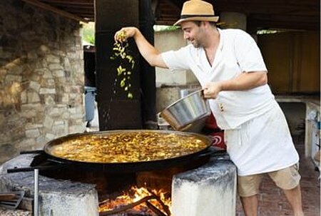 Un cuisinier préparant une grande paella sur un feu de bois dans un camping Sites et Paysages, illustrant le poste de cuisinier pour valoriser la gastronomie locale en plein air.