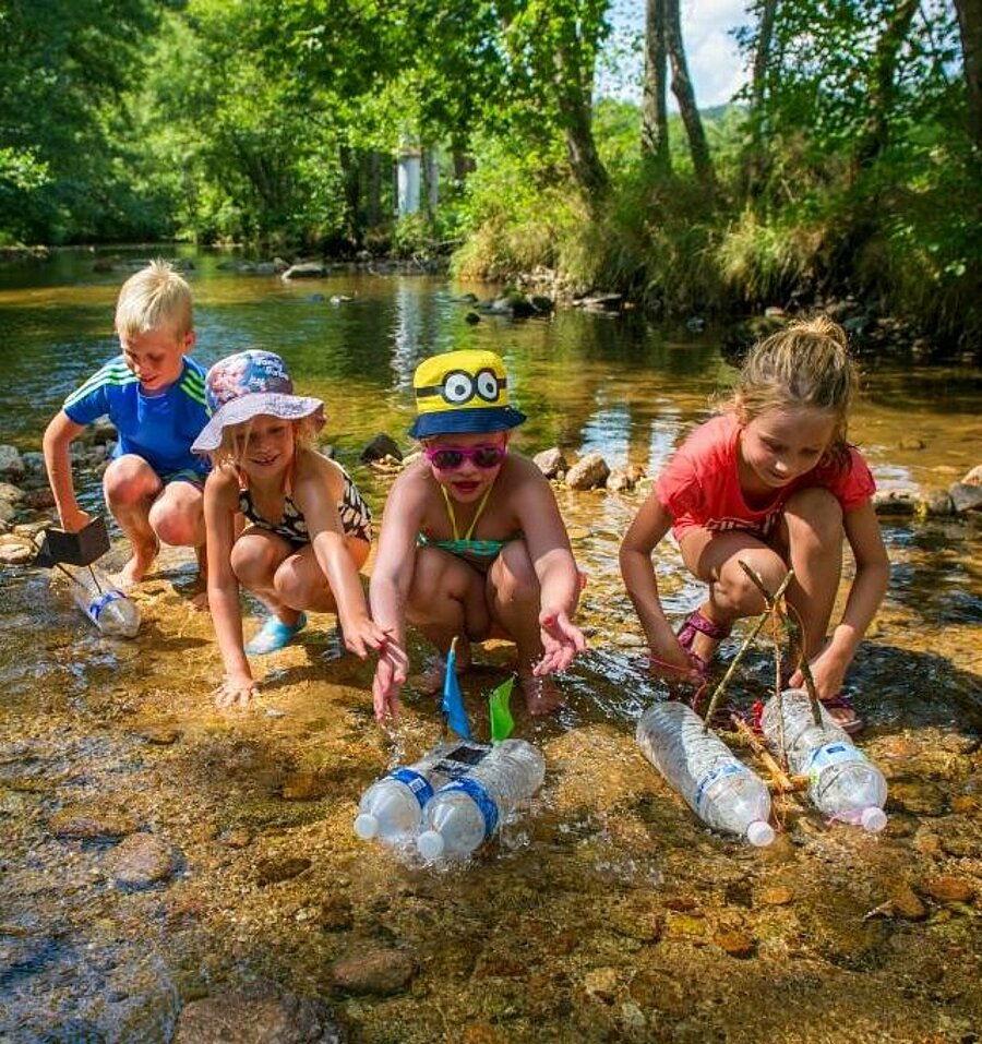 Enfants jouant dans une rivière avec des bateaux artisanaux en pleine nature, une activité ludique et conviviale en famille lors de vacances en camping en Auvergne-Rhône-Alpes.