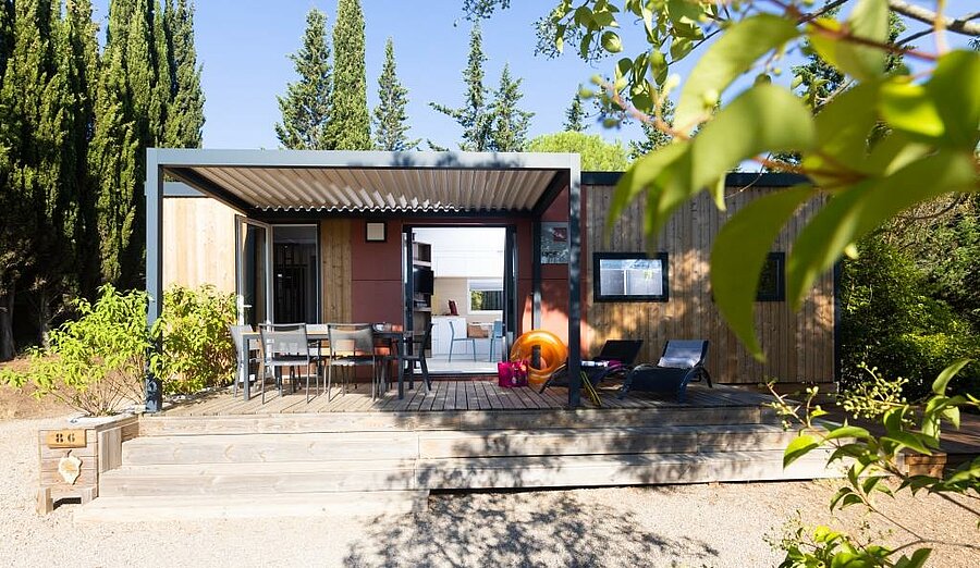 Mobil-home moderne avec grande terrasse ombragée, table, transats et végétation, dans un camping près de Carcassonne.