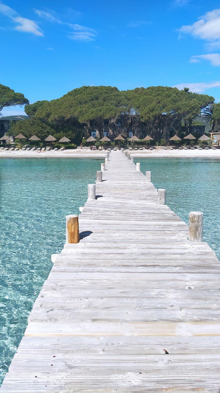 Ponton en bois menant à la plage de Palombaggia, aux eaux turquoise et cristallines en Corse, entourée de verdure et de pins parasols, parfait pour un séjour en camping.