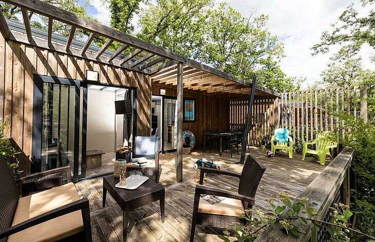 Hébergement avec terrasse semi-ombragée Terrasse en bois semi-ombragée d’un hébergement de camping entouré d’arbres.