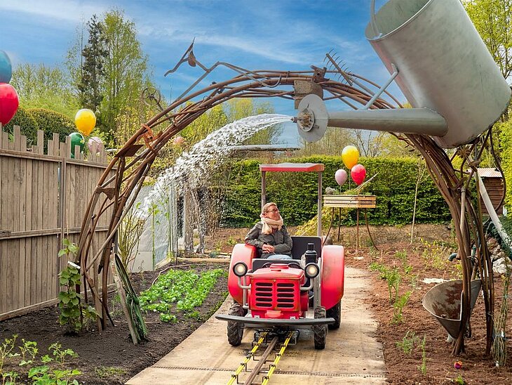 Les Tractocabines de Cléamolette © Terra Botanica Une visiteuse à bord d'un tracteur rouge passe sous un arrosoir géant qui déverse de l'eau, au milieu d'un potager.