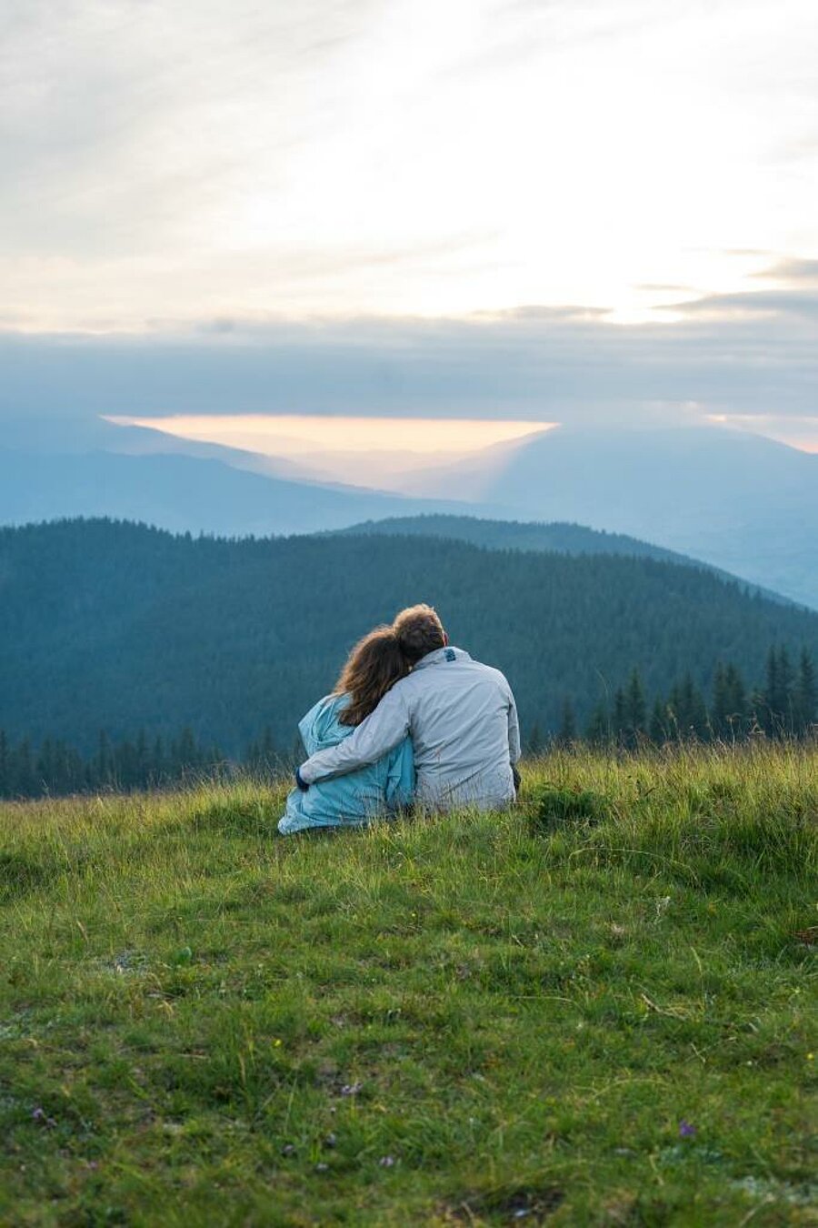 Couple enlacé assis dans l’herbe face aux montagnes au coucher du soleil, cadre paisible idéal pour un week end couple en camping nature.