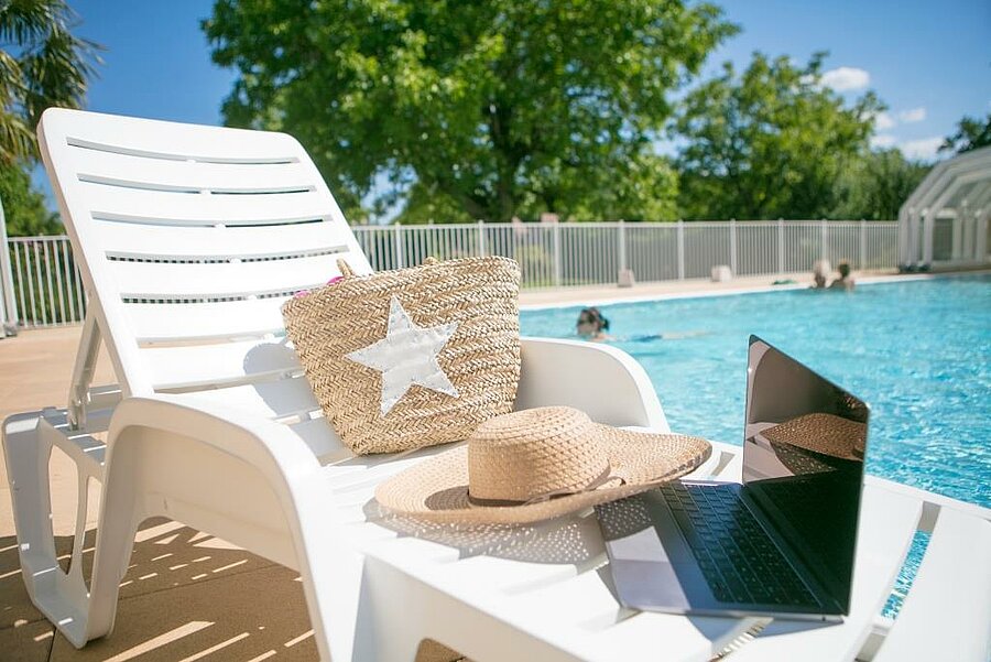 Transat au bord d’une piscine avec chapeau, panier et ordinateur portable, symbole d’un séjour relaxant et connecté dans un camping pas cher au meilleur prix garanti.