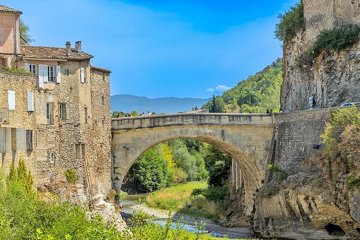 Le pont romain sur l’Ouvèze Pont romain de Vaison-la-Romaine au-dessus de l’Ouvèze, entre maisons anciennes, falaise et reliefs en arrière-plan.
