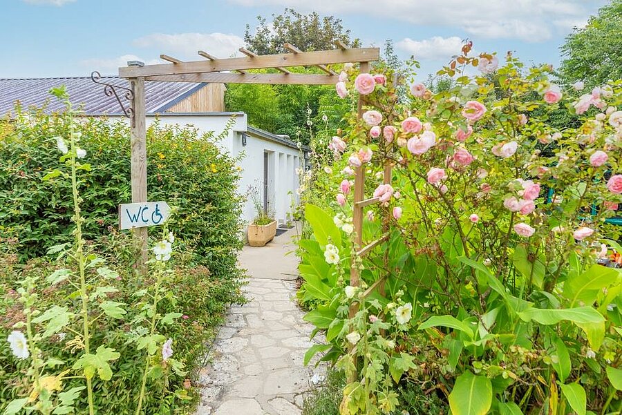 Accès fleuri aux sanitaires accessibles près d’un emplacement de camping pour caravane, dans un environnement soigné et naturel.