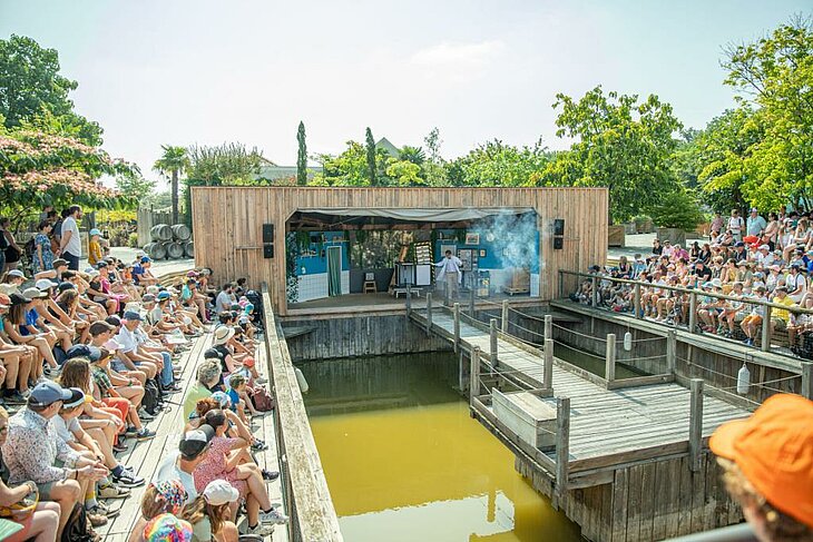Le labo magique, spectacle en plein air © Terra Botanica Des familles assistent à un spectacle sur une scène en bois au-dessus de l'eau, dans le parc Terra Botanica à Angers.