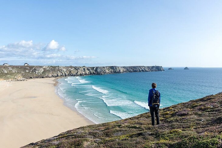 Randonneur observant une grande plage et l’océan depuis la côte sauvage du Finistère, activité nature emblématique du Finistère Tourisme.