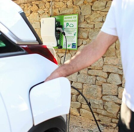 Recharge d’une voiture électrique sur une borne électrique installée dans un camping, avec un mur en pierres en arrière-plan.