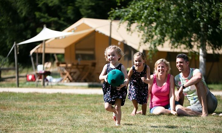 Petite fille jouant au ballon devant sa famille souriante dans un camping nature, symbole du bonheur simple des vacances anticipées.