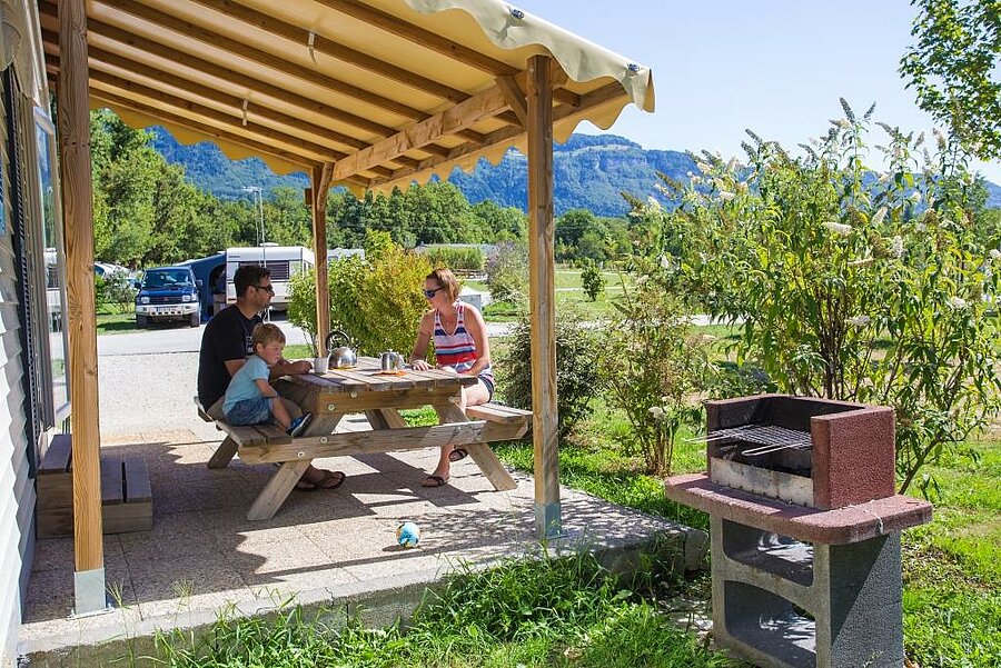 Famille en terrasse devant un mobil-home tout équipé avec barbecue dans un camping proche d’Annecy, face aux montagnes.