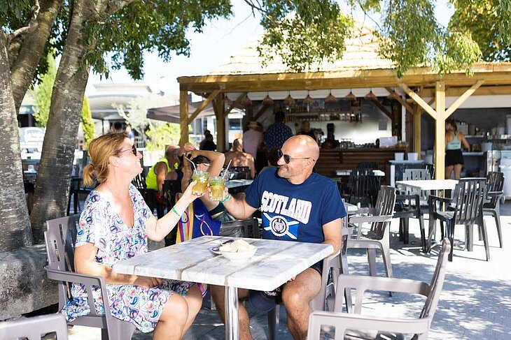 Couple en terrasse au bar du camping en early booking Couple attablé en terrasse au bar du camping, trinquant sous les arbres, scène conviviale illustrant l’early booking couple.