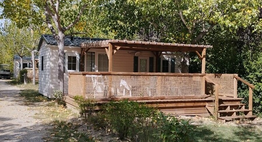 Mobil-home avec terrasse couverte en bois et salon de jardin, installé sous les arbres au Domaine de l’Écluse, dans la Drôme.