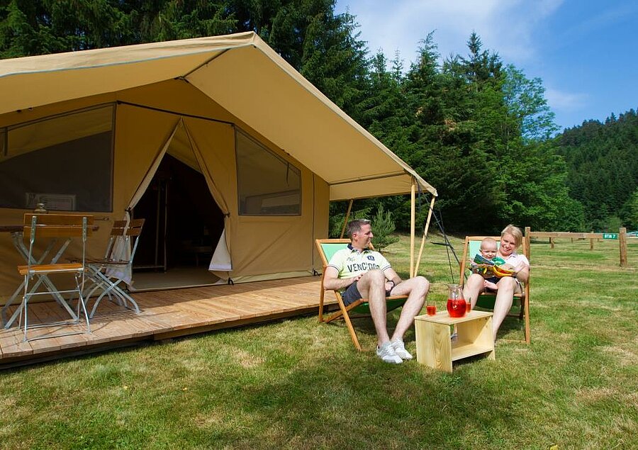 Famille détendue devant une tente lodge avec terrasse en bois dans un camping ANWB, hébergement confortable au cœur de la nature.
