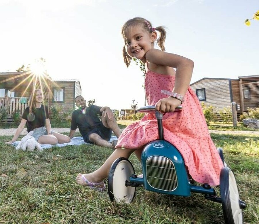 Petite fille souriante sur un porteur devant un mobil-home, ses parents détendus au soleil, image d’un séjour nature en famille avec l’offre Camping Early Booking “Un Bonheur d’Avance”.