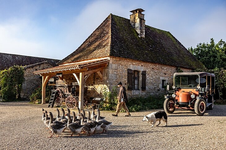 Scène au Village du Bournat avec maison en pierre, oies, chien, véhicule ancien et personnage en costume d’époque.