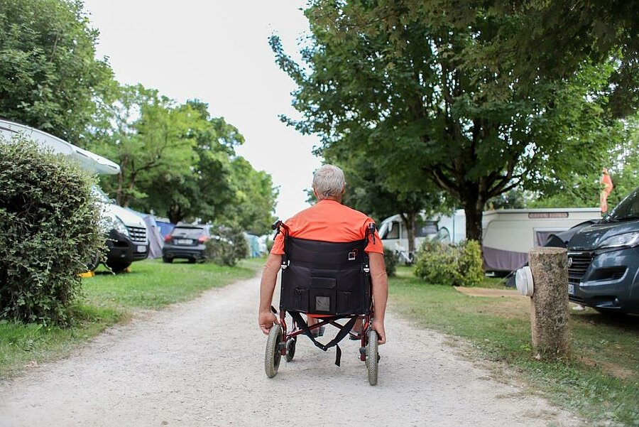 Vacancier en fauteuil roulant circulant sur une allée de camping arborée, illustrant un logement PMR avec services adaptés et confortables.