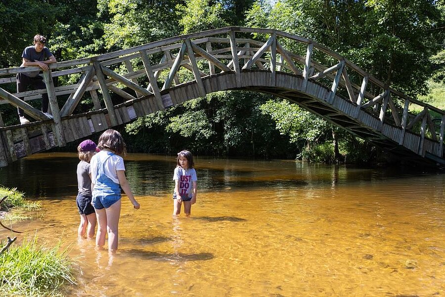 Enfants jouant dans une rivière peu profonde sous un pont en bois, dans un camping pas cher et nature idéal pour des vacances familiales au bord de l'eau