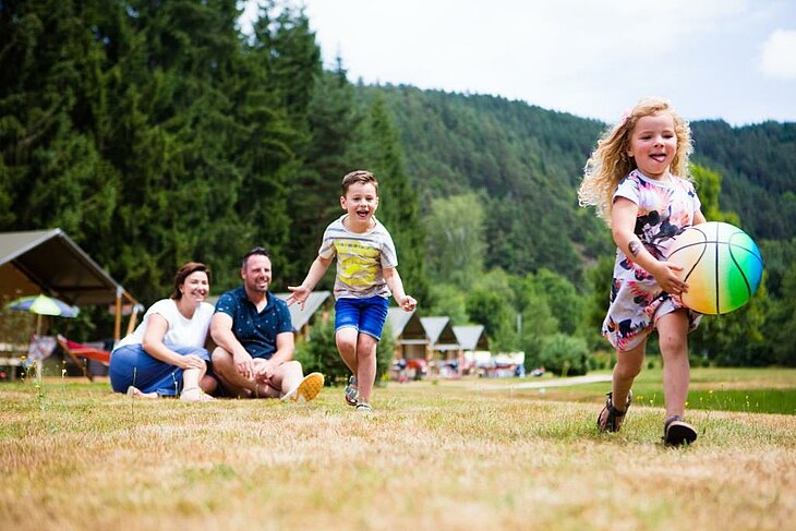 Enfants jouant au ballon sur l’herbe pendant les vacances en camping nature, parents souriants en arrière-plan.