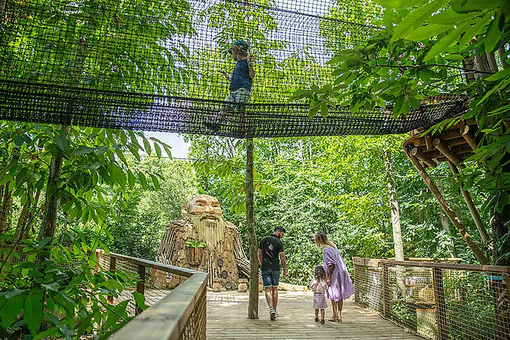Le Parcours des géants © Terra Botanica Un enfant traverse un filet suspendu au-dessus d'une passerelle en bois, près d'une sculpture de géant à Terra Botanica.