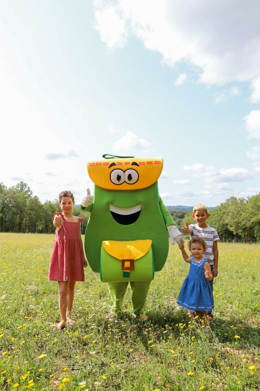 Mascotte PAKO et enfants dans un pré fleuri, ambiance camping dernière minute août en pleine nature ensoleillée.