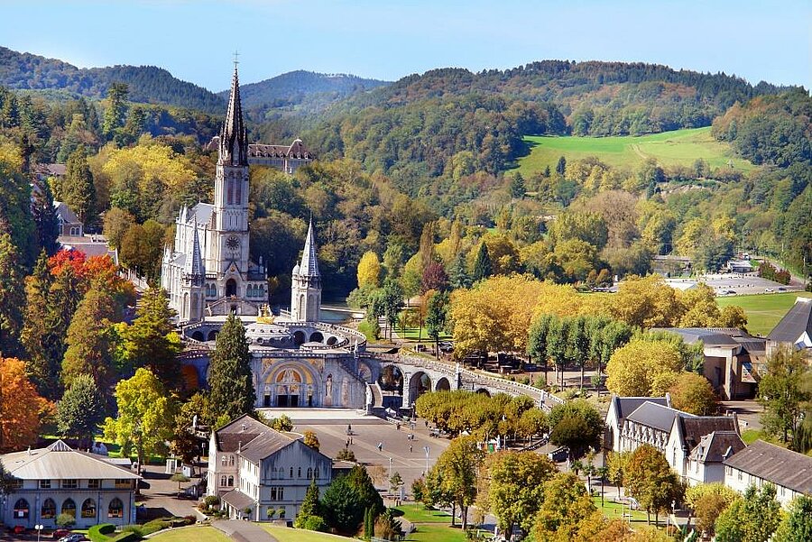 Vue aérienne sur le sanctuaire et la basilique de Lourdes entourés de forêts et montagnes, près d’un camping dans les Pyrénées.