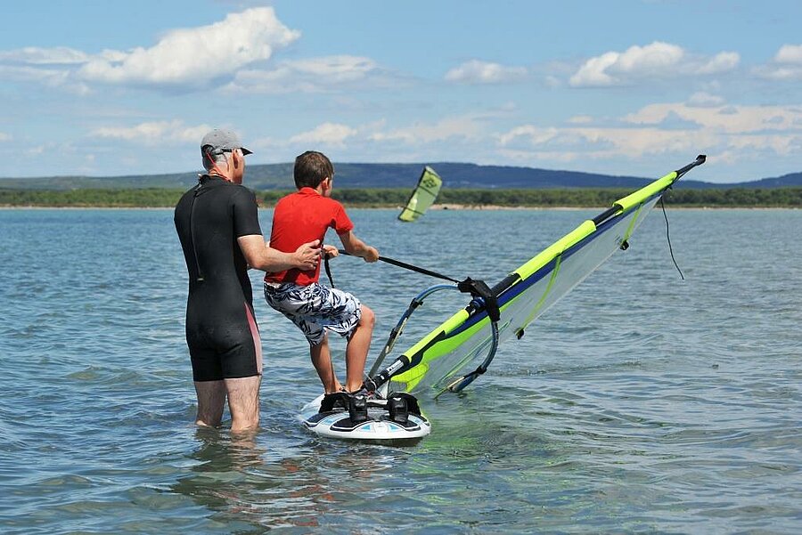 Enfant initié à la planche à voile avec un adulte dans un lac calme, activité idéale en camping planche à voile en famille.