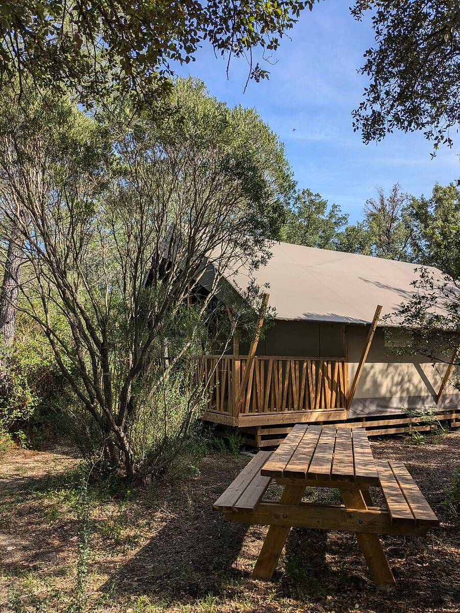 Tente lodge en bois et toile avec terrasse, entourée de verdure et équipée d’une table de pique-nique dans un camping en Corse, idéale pour un séjour confortable en pleine nature.