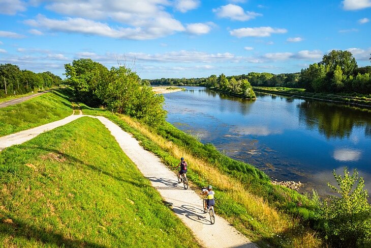 Balade à vélo le long de la Loire Deux cyclistes parcourent une piste aménagée le long de la Loire, entre prairies verdoyantes, arbres et berges naturelles.