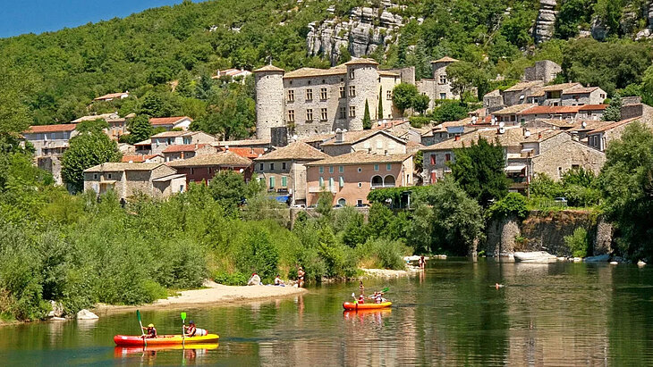 Vue sur Vogüé, ses maisons en pierre et son château dominant l’Ardèche, avec canoës et baignade au pied du village.