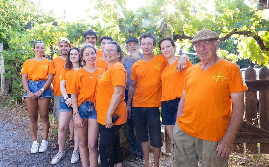 Photo de l’équipe du camping La Pinède près de Carcassonne, souriante et vêtue de t-shirts orange sous une pergola de vigne.