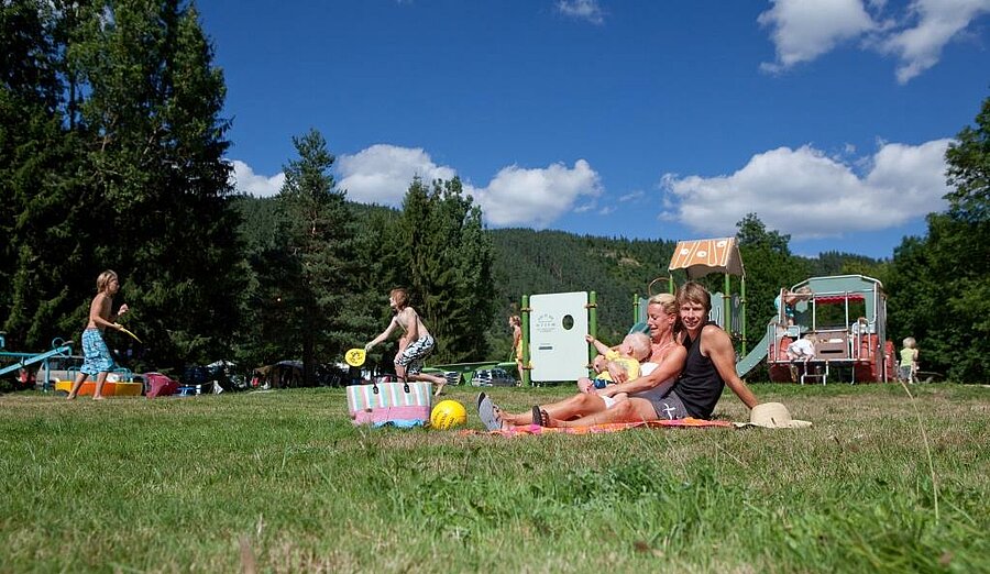 Famille détendue sur la pelouse près d’une aire de jeux dans un camping ANWB, ambiance conviviale et nature pour des vacances réussies.