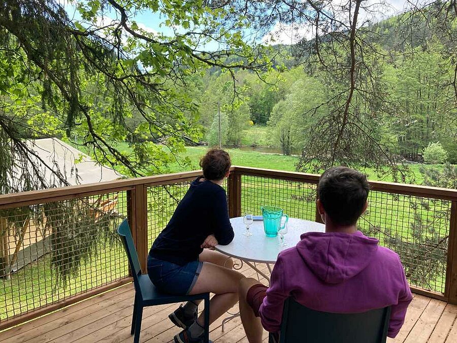 Deux personnes assises sur une terrasse en bois, face à une prairie et une forêt verdoyante, dans un camping du Massif central.