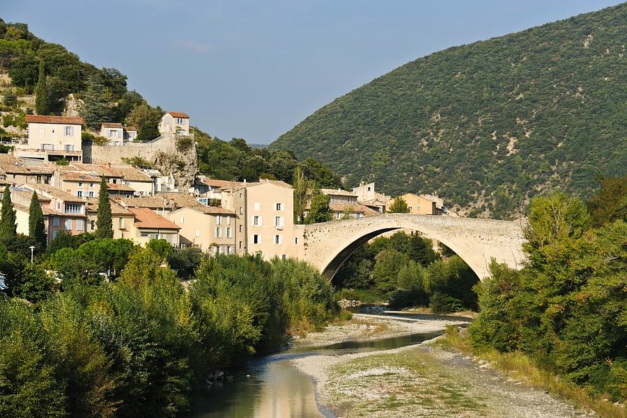 Vue du village de Nyons et de son pont médiéval sur la rivière, entouré de collines verdoyantes de la Drôme provençale.