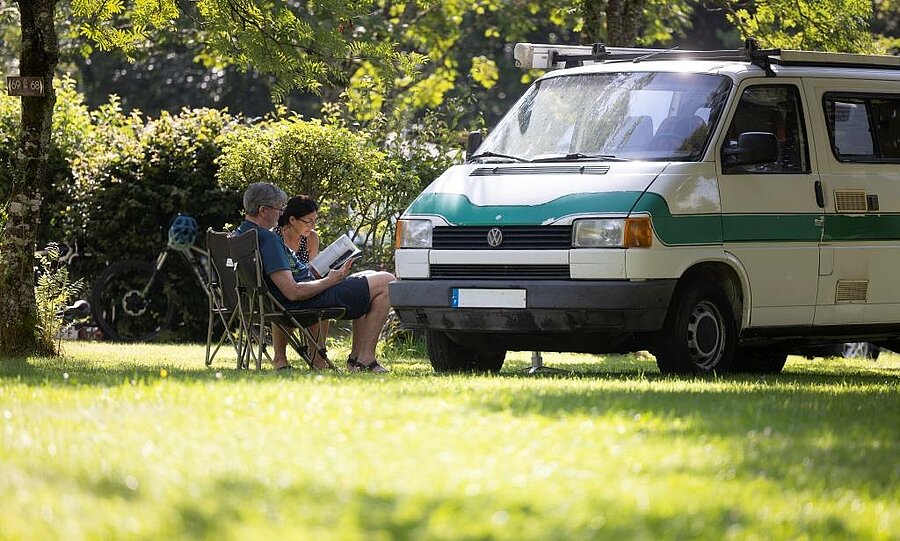Couple installé devant un camping van sur un emplacement van arboré, illustrant la liberté et la simplicité de la vanlife en pleine nature.