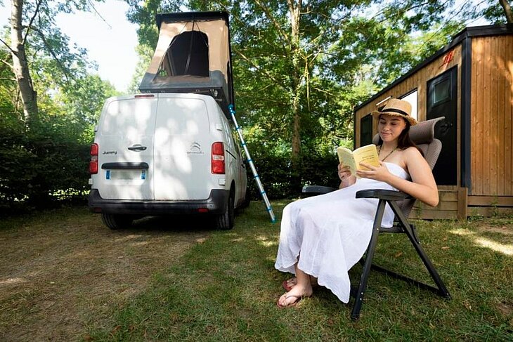 Séjour en van sur un emplacement ombragé Vacancière lisant à l’ombre près d’un van aménagé avec toit relevable dans un camping