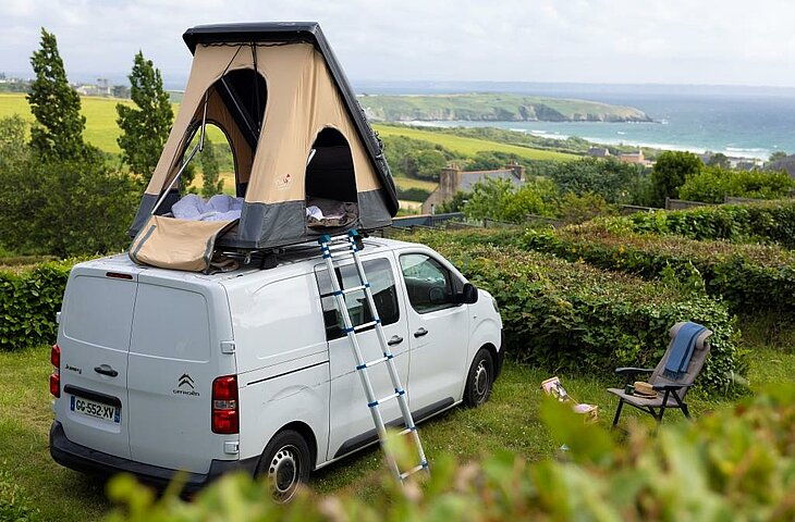Van avec tente de toit installé sur un emplacement verdoyant face à la mer, idéal pour un séjour nature et confort.