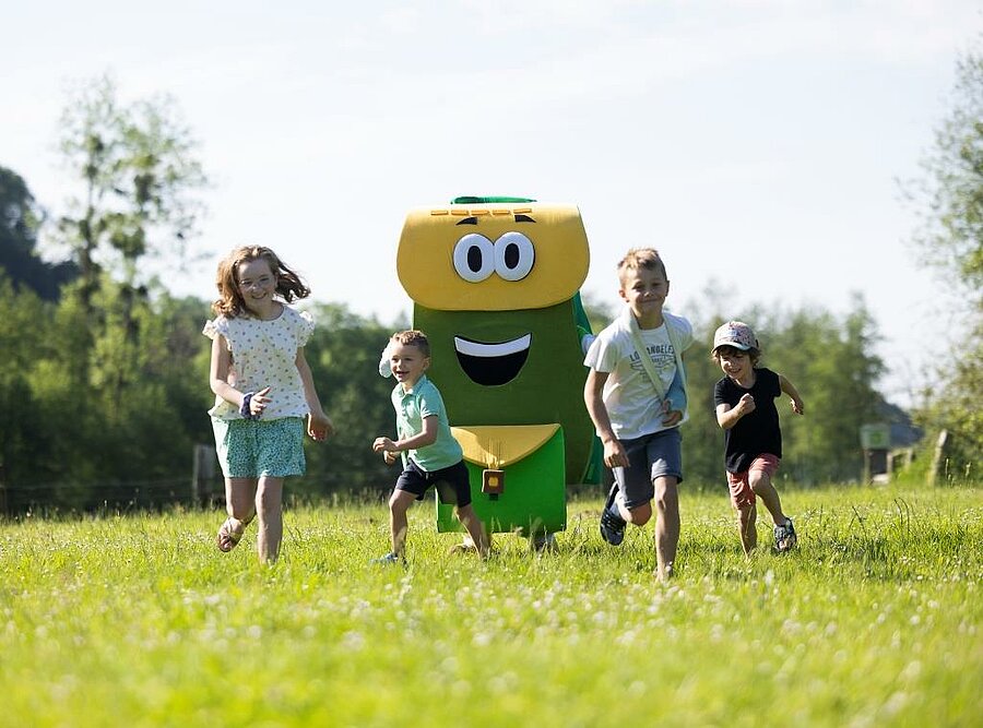 Enfants courant dans l’herbe avec la mascotte PAKO, ambiance vacances en famille conviviale en pleine nature.