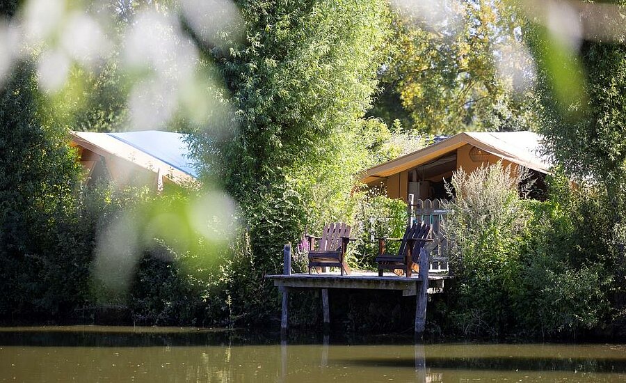 Tente lodge et terrasse en bois avec fauteuils, au bord d’un étang calme dans un camping nature des Pays de la Loire.