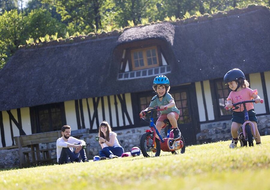 Deux enfants faisant du vélo devant un hébergement typique normand, pendant que leurs parents profitent d’un moment de détente dans un camping près de Honfleur.